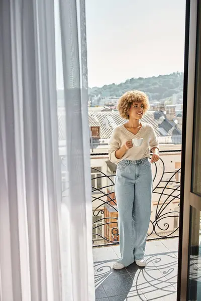 Joyeuse femme afro-américaine frisée debout avec une tasse de café sur le balcon de l'hôtel, vue sur la ville — Photo de stock