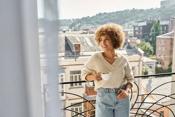 Soñadora mujer afroamericana rizada de pie con taza de café en el balcón del hotel, vista a la ciudad - foto de stock