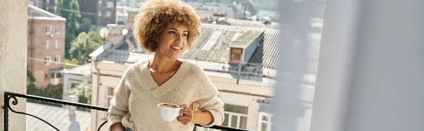 Mujer afroamericana joven soñadora de pie con taza de café en el balcón del hotel, pancarta de viaje - foto de stock