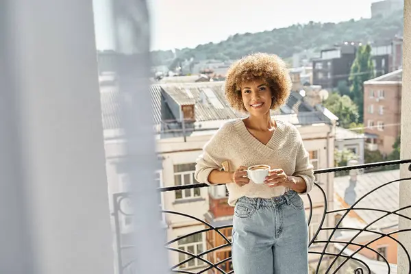 Feliz afro-americano encaracolado mulher de pé com xícara de café na varanda do hotel, vista da cidade — Fotografia de Stock