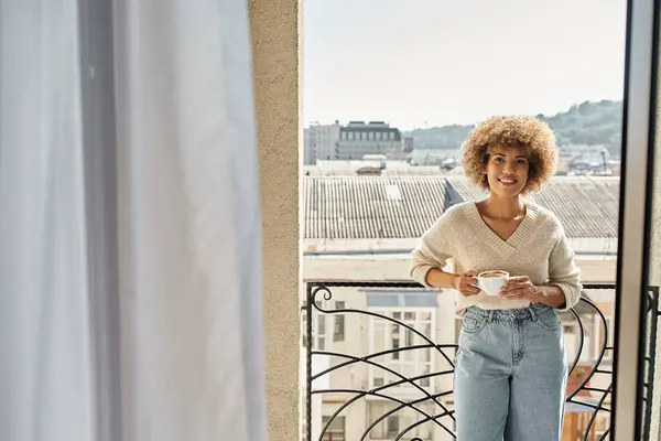 Smiling curly african american woman standing with cup of coffee on hotel balcony, city view — Stock Photo