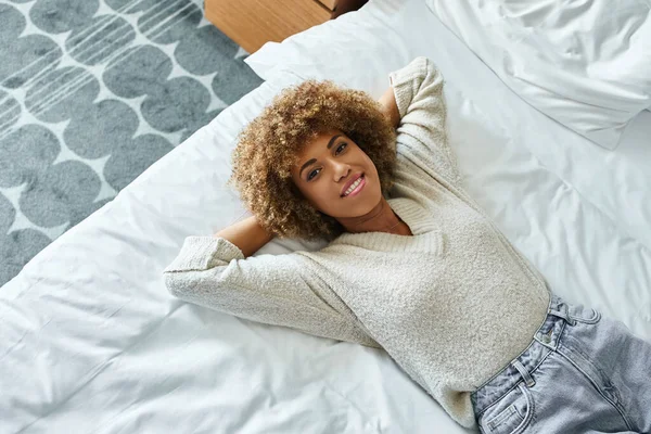 Vue de dessus de la femme afro-américaine détendue souriante et couchée sur le lit dans la chambre d'hôtel, voyageur seul — Photo de stock
