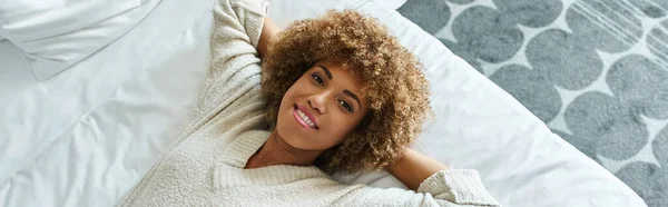 Striscione di rilassata donna afroamericana sorridente e sdraiata sul letto in camera d'albergo, viaggiatore solitario — Foto stock
