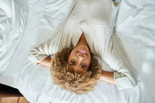 Vista superior da mulher americana africana relaxada sorrindo e deitada na cama no quarto de hotel, de cabeça para baixo — Fotografia de Stock
