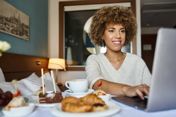 Joyeuse femme afro-américaine utilisant son ordinateur portable près du petit déjeuner à l'hôtel, service en chambre et commodité — Photo de stock