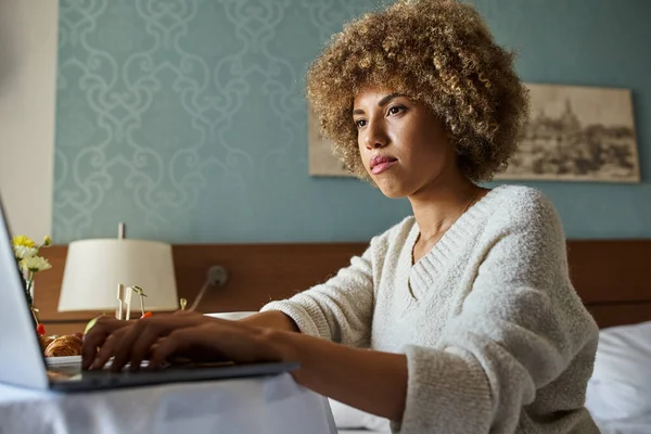 Focalisé jeune femme afro-américaine travaillant à distance sur son ordinateur portable dans une chambre d'hôtel, freelance — Photo de stock
