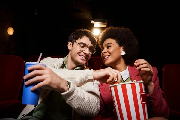Cheerful young multicultural couple in retro clothes enjoying date at cinema and sharing popcorn — Stock Photo