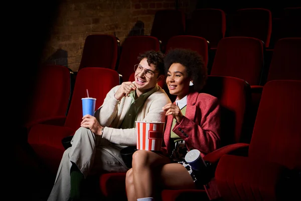 Счастливая молодая мультикультурная пара в стильных нарядах, смотрящая кино на свидании в кино, День Святого Валентина — стоковое фото