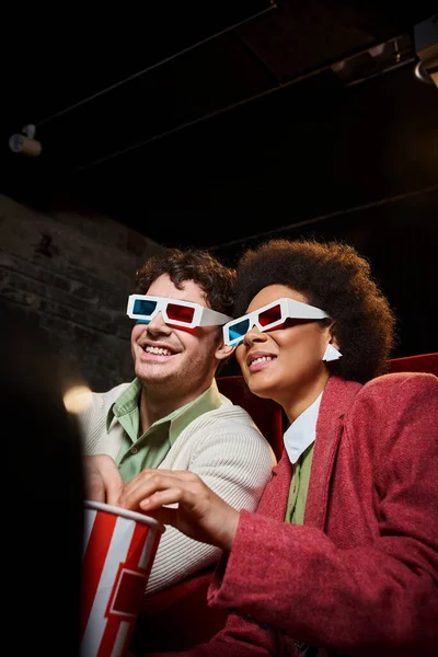 Cheerful multiethnic couple with retro 3d glasses and popcorn watching movie on date at cinema — Stock Photo