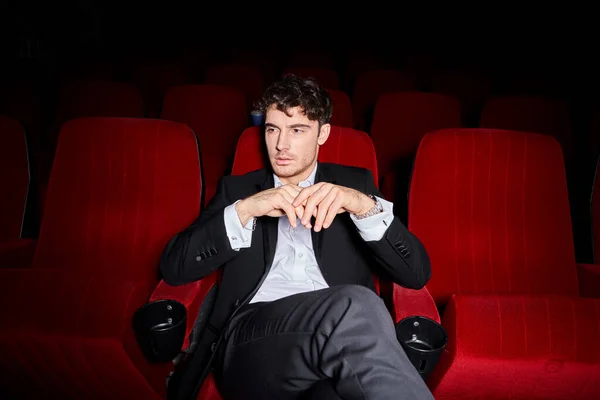 Bom olhar modelo masculino elegante com estilo dapper sentado em cadeiras de cinema vermelho e olhando para longe — Fotografia de Stock