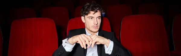 Hombre elegante guapo con estilo elegante sentado en sillas de cine rojas y mirando hacia otro lado, pancarta - foto de stock