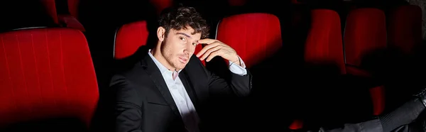 Modelo masculino elegante em terno chique preto sentado na cadeira de cinema vermelho com a mão perto do rosto, banner — Fotografia de Stock