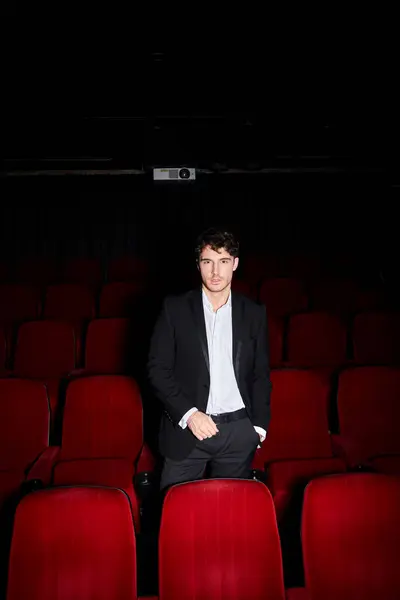 Young appealing man in elegant black suit posing near red cinema chairs and looking at camera — Stock Photo