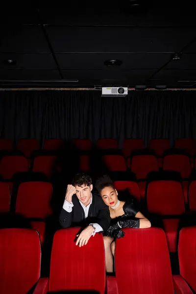 Ein attraktives multikulturelles Stilpaar in schwarzen Abendkleidern sitzt auf roten Stühlen im Kino — Stockfoto
