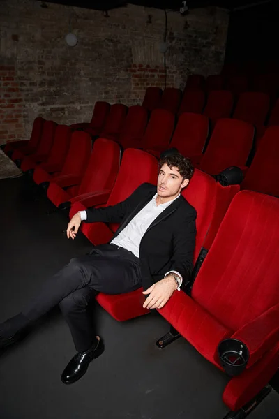 Good looking male model in elegant stylish suit sitting on red chair at cinema, looking at camera — Stock Photo