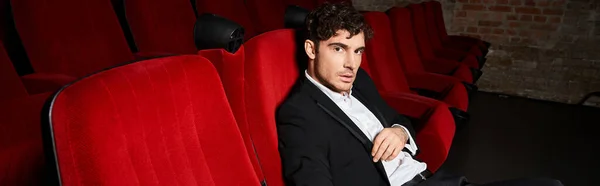 Young appealing man in chic black suit sitting on red chairs at cinema and looking at camera, banner — Stock Photo