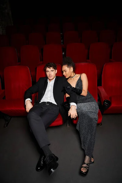 Привлекательная многорасовая пара в вечерних нарядах, сидящая на красных стульях кинотеатра, День святого Валентина — стоковое фото