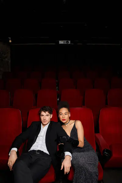 Young attractive multicultural couple in elegant attires sitting on red cinema chairs on date — Stock Photo
