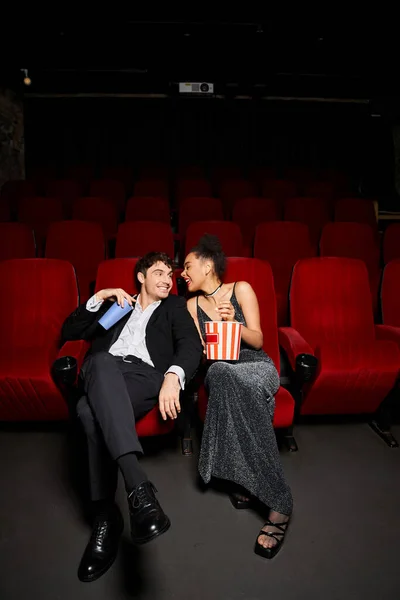 Happy young diverse couple in evening attires holding popcorn and soda and smiling at each other — Stock Photo