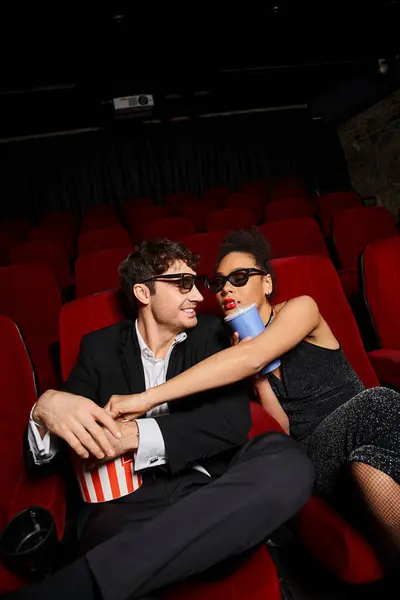 Alegre pareja multirracial con gafas 3d en trajes elegantes compartiendo refrescos y palomitas de maíz en la fecha en el cine - foto de stock