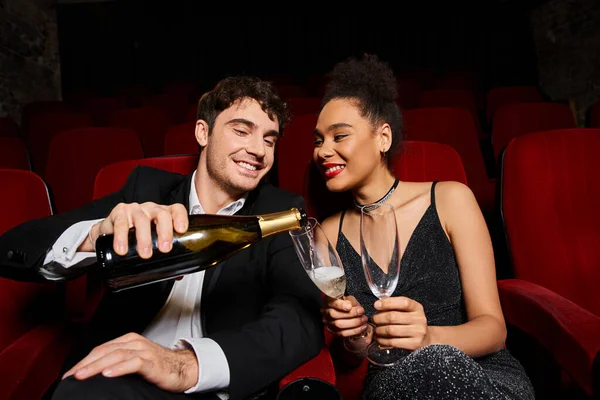 Feliz pareja interracial disfrutando de una cita con champán en el cine y sonriendo felizmente, el día de San Valentín - foto de stock