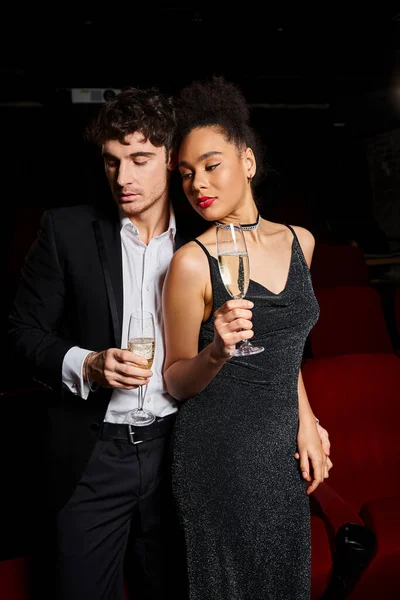 Atraente jovem casal multiétnico em trajes elegantes pretos posando com champanhe e olhando para longe — Fotografia de Stock
