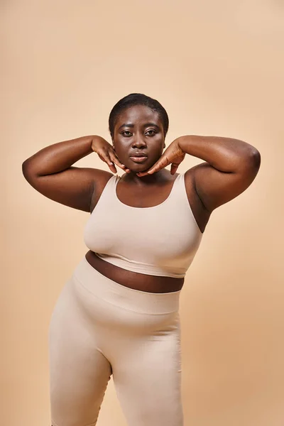 Plus size African American woman in beige underwear posing with hands near face on beige background — Stock Photo