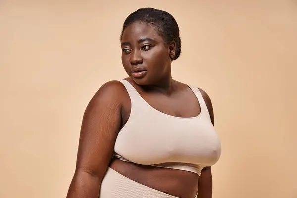 Plus size African American woman in beige undergarments posing thoughtfully on beige background — Stock Photo