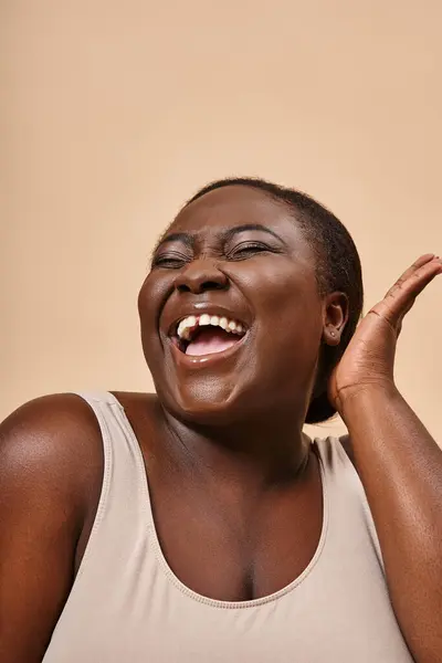 Alegre más tamaño africano americano mujer sonriendo con la mano cerca de su cara sobre fondo beige - foto de stock
