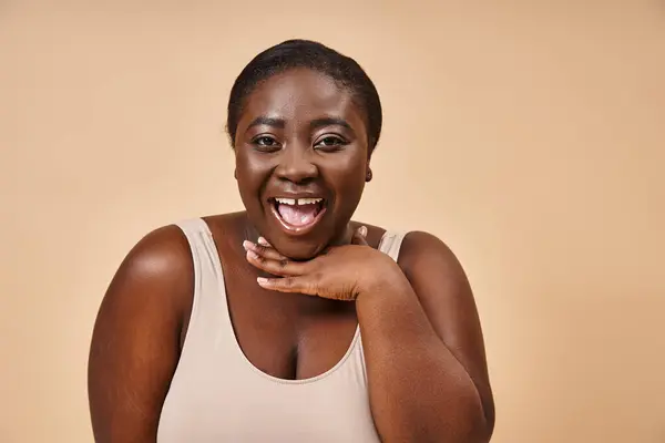 Excited plus size african american woman smiling with hand near her face on beige background — Stock Photo