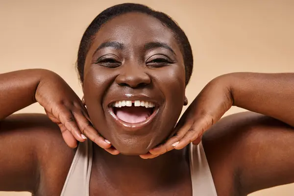 Cheerful plus size african american woman smiling with hands near her face on beige background — Stock Photo