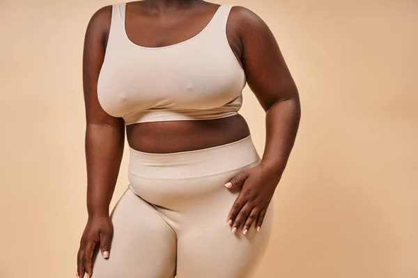 Обрізаний плюс розмір афроамериканської жінки в нижній білизні позує на бежевому фоні, позитивне тіло — стокове фото