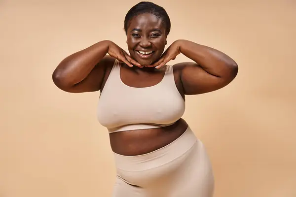 Allegro plus size donna africana americana sorridente con le mani vicino al viso su sfondo beige — Foto stock