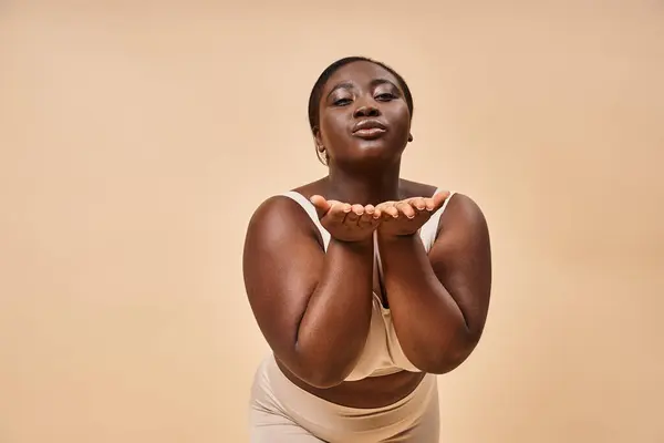 Joven más tamaño afroamericana mujer en ropa interior beige soplado aire beso contra juego telón de fondo - foto de stock