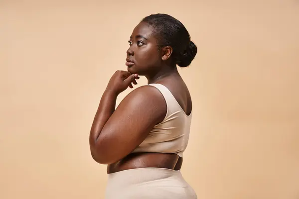 Pensivo plus size afro-americano mulher em roupa interior bege posando contra o pano de fundo correspondente — Fotografia de Stock
