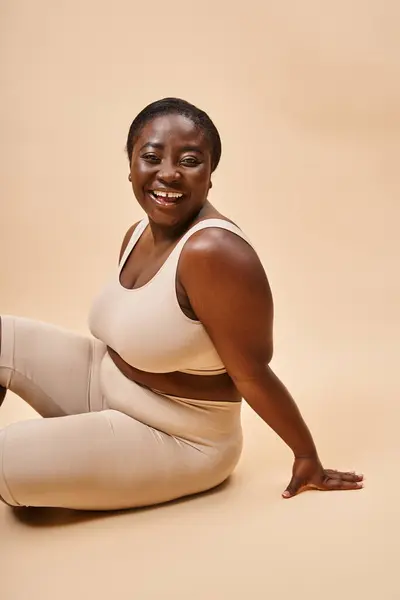 Alegre más tamaño afroamericano joven mujer en ropa interior beige posando contra el telón de fondo a juego - foto de stock