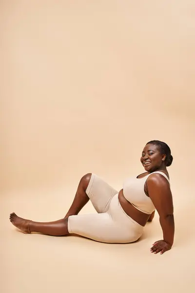 Heureux plus la taille afro-américaine jeune femme en sous-vêtements beige posant sur fond correspondant — Photo de stock