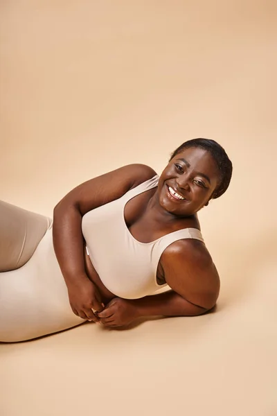 Heureux plus la taille afro-américaine jeune femme en lingerie beige inclinable sur fond assorti — Photo de stock