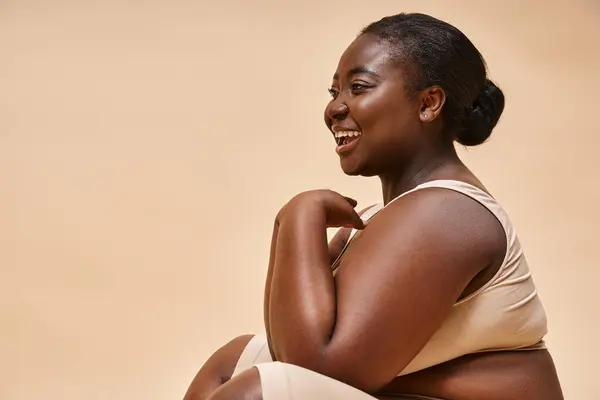 Modelo afroamericano de tamaño grande riéndose y sentado sobre fondo beige, cuerpo positivo - foto de stock