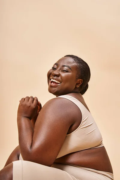 Plus size african american model in beige lingerie laughing and reclining against matching backdrop — Stock Photo