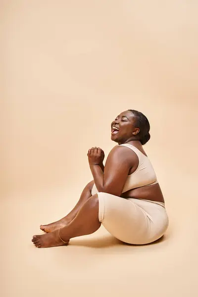 Plus size afrikanisch amerikanische Frau lachend und sitzend vor beigem Hintergrund, Körper positiv — Stockfoto