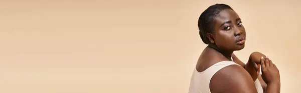 Banner positivo para el cuerpo, modelo afroamericano de talla grande en ropa interior posando sobre fondo beige - foto de stock