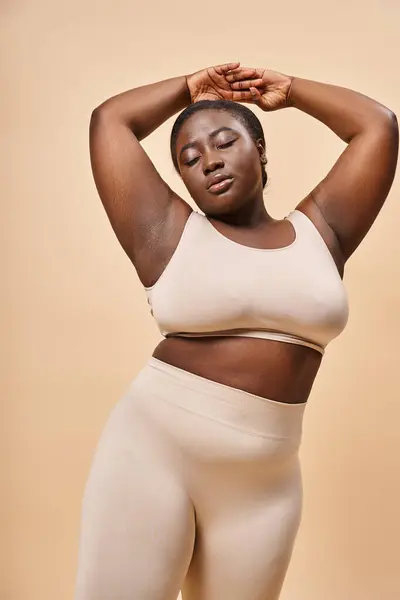 Plus la taille femme afro-américaine en sous-vêtements debout avec les mains levées sur fond beige — Photo de stock