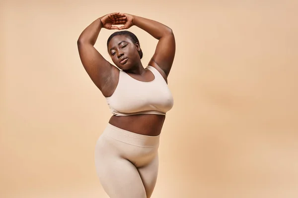 Más tamaño mujer afroamericana en ropa interior posando con las manos levantadas contra el telón de fondo beige - foto de stock