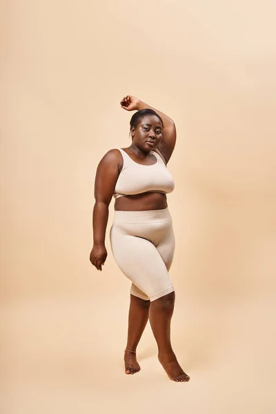 Modèle afro-américain taille plus en sous-vêtements debout avec la main levée sur fond beige — Photo de stock