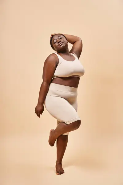 Plus size mulher americana africana em roupa interior em pé com a mão levantada contra o pano de fundo bege — Fotografia de Stock