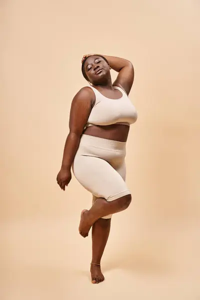 Plus size african american woman in underwear standing with raised hand against beige backdrop — Stock Photo