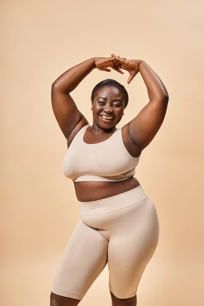 Happy plus size woman in beige underwear posing with raised hands, body positive and self esteem — Stock Photo