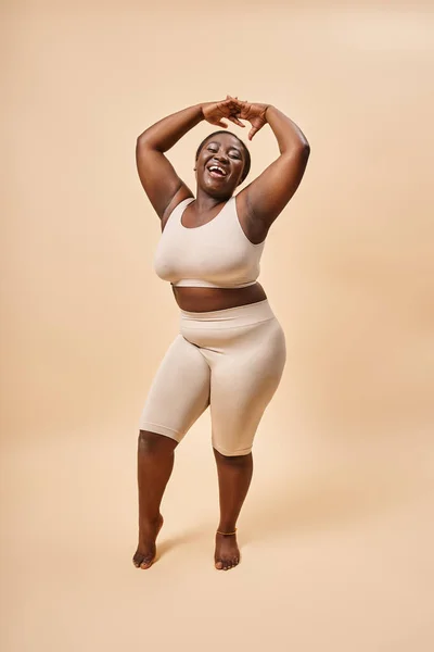 Happy plus size woman in beige underwear posing with raised hands, body positive and self esteem — Stock Photo