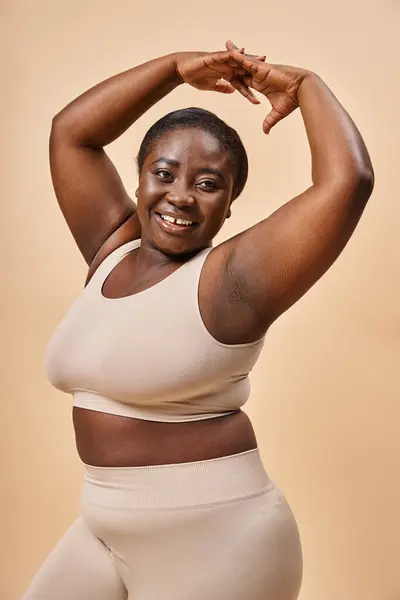 Smiling plus size woman in beige underwear posing with raised hands, body positive and self esteem — Stock Photo